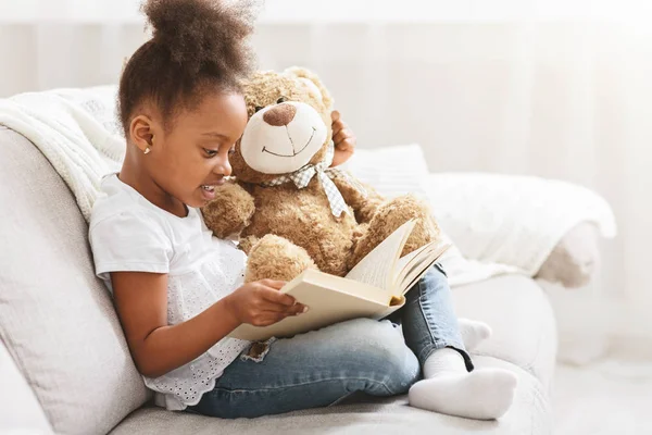 Little afro girl holding teddy bear and reading book — 图库照片
