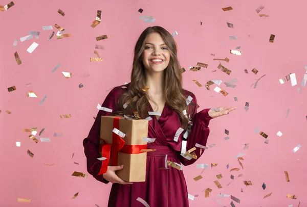 Щаслива дівчина тримає подарунок, стоячи під душем конфетті над рожевим тлом — стокове фото