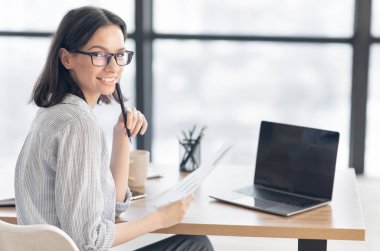 Smiling young female worker sitting with papers and laptop