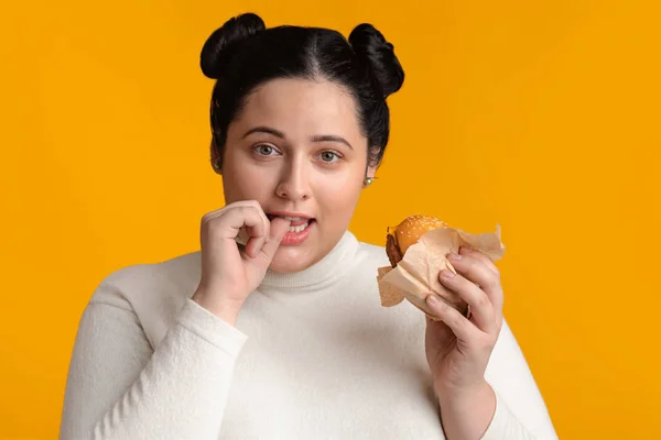 Mladá baculatá žena drží hamburger a kousá si prst — Stock fotografie