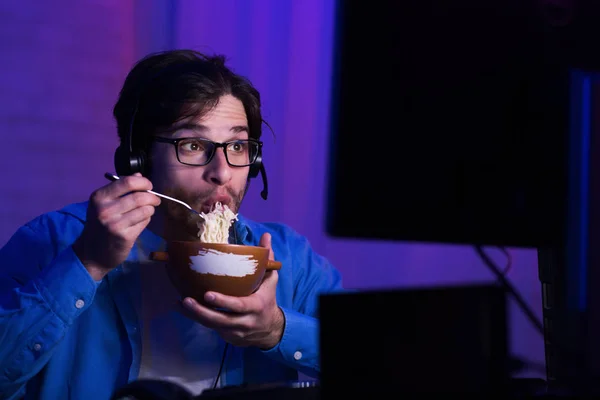 Cara jogando jogo de computador comendo sopa de macarrão em casa à noite — Fotografia de Stock