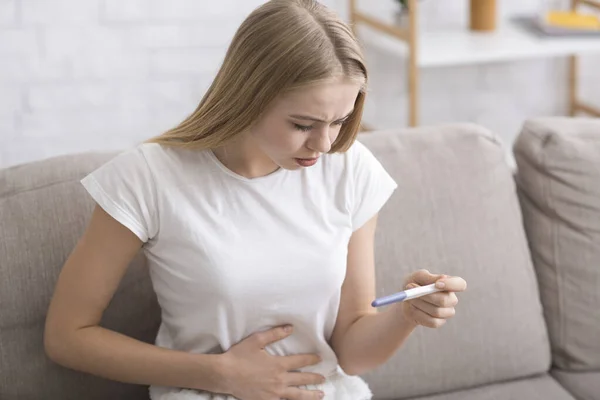 Chica joven sorprendida sosteniendo la prueba de embarazo, sentado en el sofá — Foto de Stock