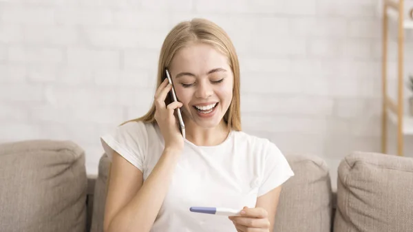 Mujer joven y feliz hablando por teléfono, sosteniendo la prueba de embarazo — Foto de Stock