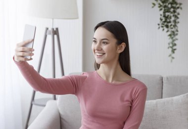 Genç, gülümseyen bir kadın cep telefonu kamerasıyla selfie çekiyor.