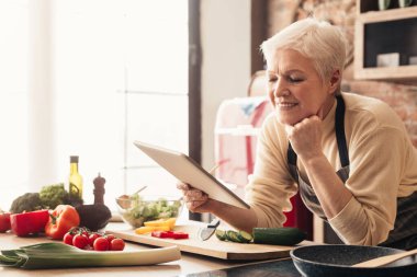 Elderly woman using digital tablet in kitchen, checking cooking recipes