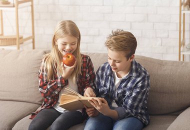 Brother And Sister Reading Book Sitting On Sofa At Home