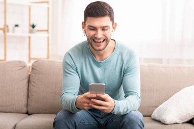 Guy In Earbuds Using Phone Sitting On Couch At Home