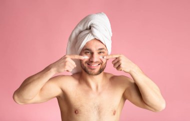 Cute young guy after a shower, white towel on head, smears cream on face