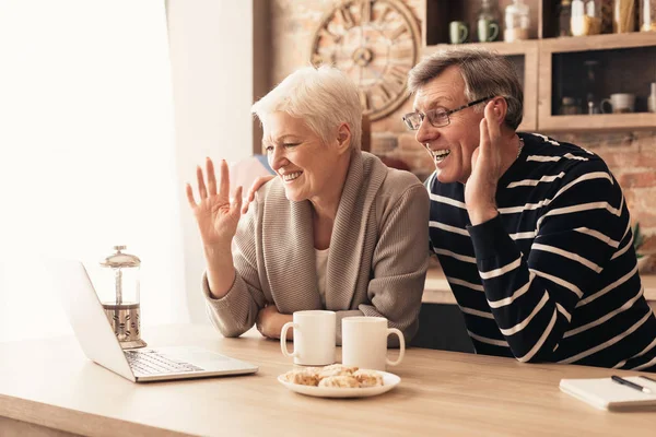 Happy senior couple looking at laptop screen in kitchen and waving — Stok fotoğraf