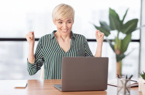 Счастливая предпринимательница, пожимающая кулаки, сидит за ноутбуком в современном офисе — стоковое фото