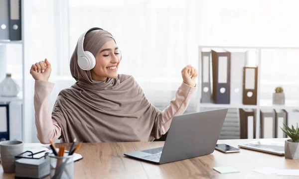 Perempuan arab yang ceria dalam musik jilbab mendengarkan musik di headphone di tempat kerja — Stok Foto