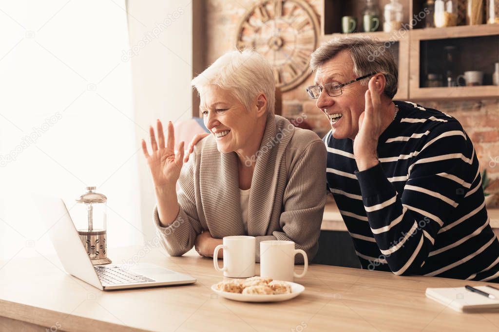 Happy senior couple looking at laptop screen in kitchen and waving