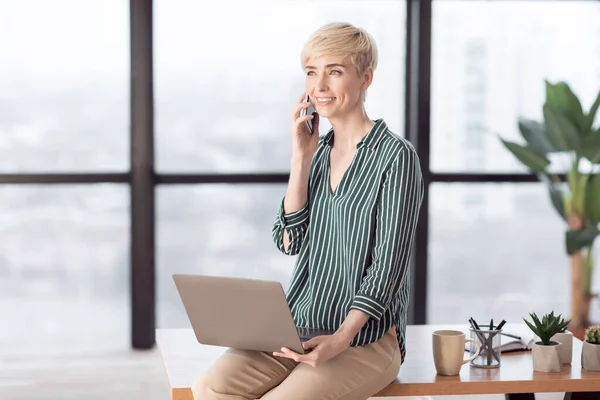 Entrepreneur Woman Talking On Cellphone Sitting On Desk In Office