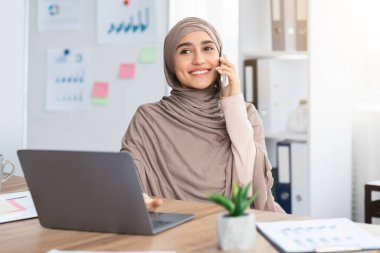 Joyful Muslim Woman Having Call While Working On Laptop In Office