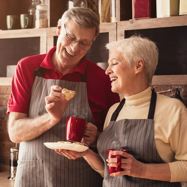 Alegre envejecido pareja comer casero galletas y beber té en cocina —  Fotos de Stock