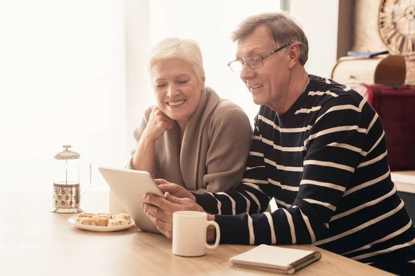 Cheerful elderly couple using digital tablet in kitchen together, reading news — Stok fotoğraf