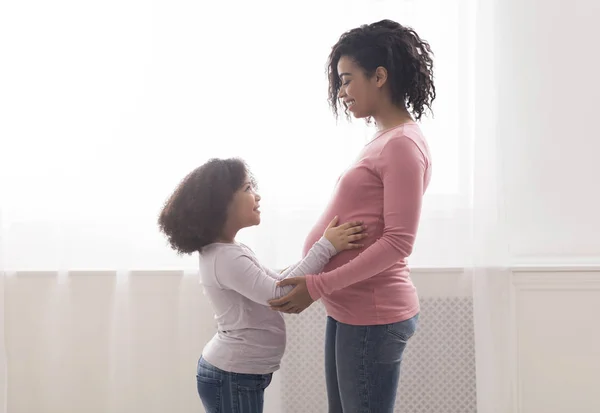 Adorable little afro daughter touching belly of her pregnant mom — Stock fotografie
