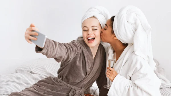 Two ecstatic women taking selfie in bathrobes, drinking champagne — Stok fotoğraf