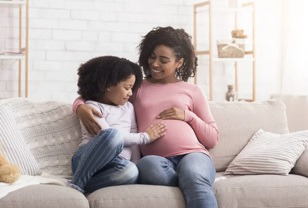 Допитлива маленька дочка торкається вагітної мами животик, сидячи на дивані вдома, порожній простір — стокове фото