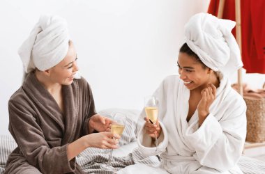 Multiracial girlfriends in bathrobes celebrating weekend, drinking champagne