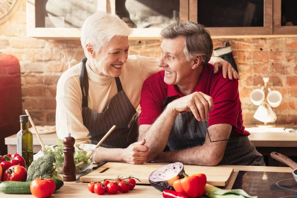 Happy elderly couple embracing in kitchen while cooking dinner together — Stok fotoğraf