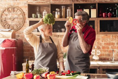 Elderly couple having fun in kitchen, playing with vegetables clipart