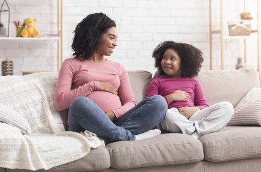 Pregnant afro mom and her daughter touching bellies, sitting on couch