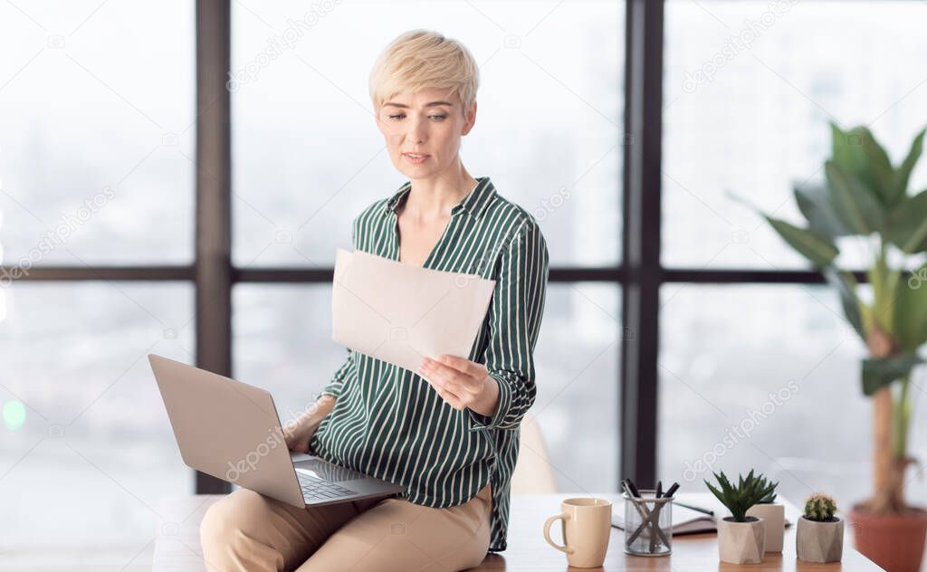 Lady With Laptop Reading Papers Sitting On Desk In Office