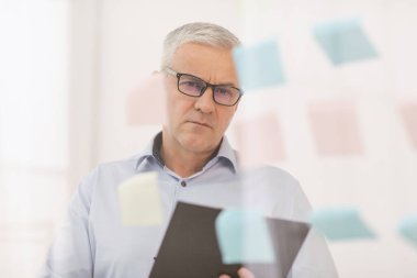 Pensive mature manager looking at sticky notes on glass