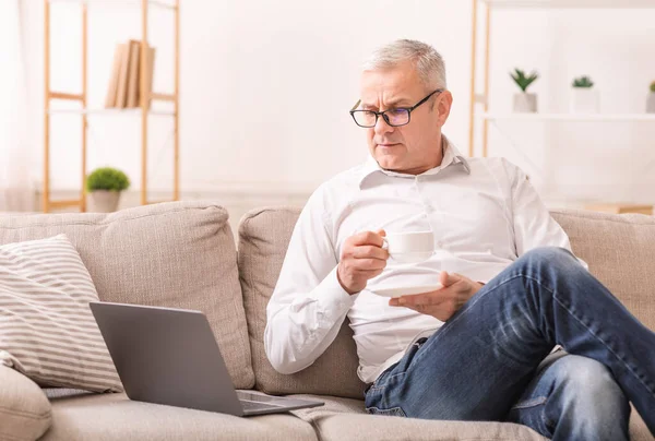 Anciano mirando a la computadora portátil y tomando té — Foto de Stock