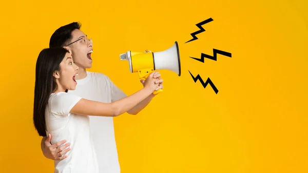 Young asian couple shouting in megaphone over yellow background — Stok fotoğraf