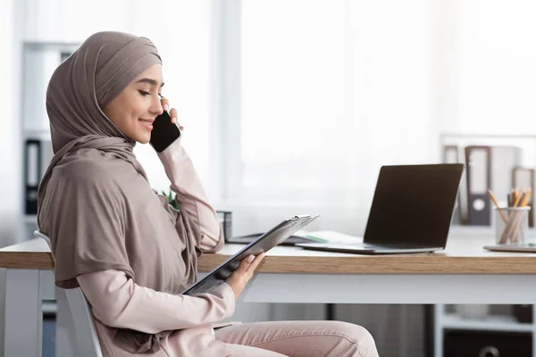 Muslim businesswoman talking on cellphone, holding clipboard, checking documentation in office — Stockfoto