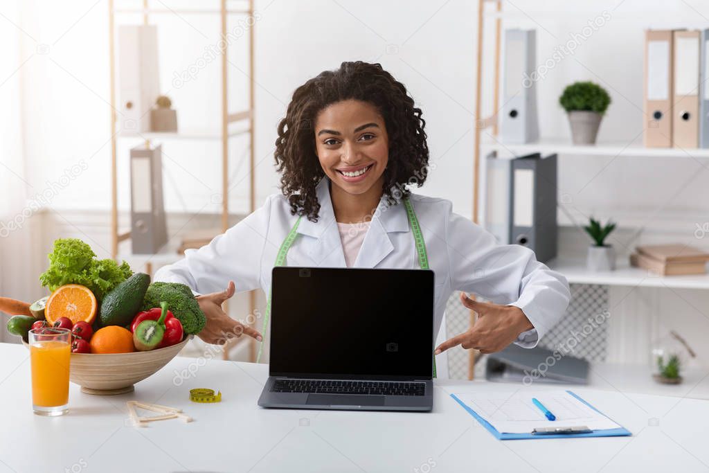 Cheerful black woman dietologist pointing at empty laptop screen