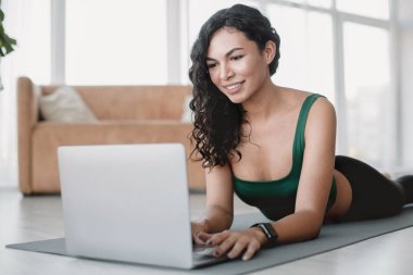 Smiling Hispanic woman with laptop on yoga mat indoors