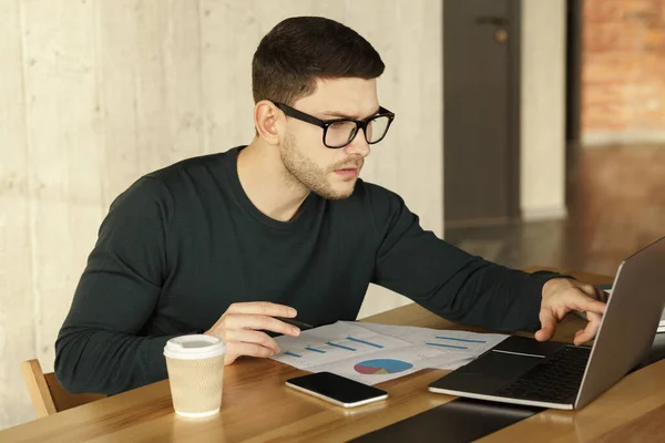 Занятой молодой человек работает с картами, сидя на рабочем месте в помещении — стоковое фото