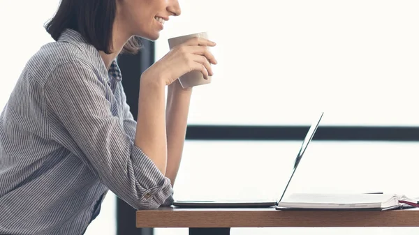 Jonge vrouw met laptop zittend aan het bureau door het raam — Stockfoto