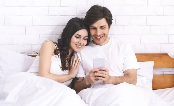 Jóvenes cónyuges felices en red en el teléfono celular en la cama — Foto de Stock