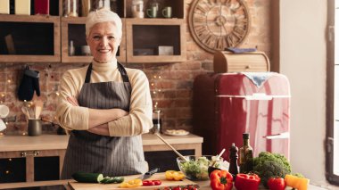 Mutfakta kollarını kavuşturmuş yaşlı kadın pozu veriyor, kameraya bakıyor.