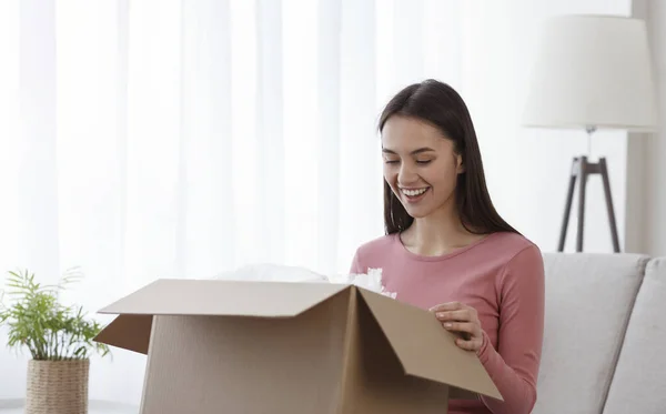 Opgewonden duizendjarige vrouw zittend op de bank thuis openen kartonnen doos — Stockfoto