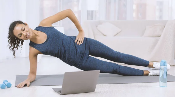 Leende latinsk kvinna gör sidoplanka motion medan du tittar på träningsvideo på bärbar dator hemma — Stockfoto