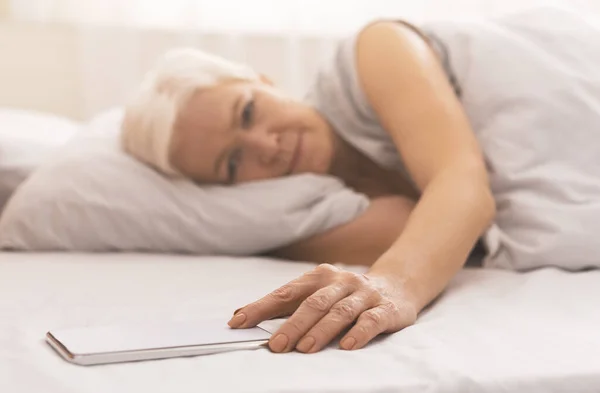 Mujer mayor cansada en la cama revisando la hora en su teléfono — Foto de Stock