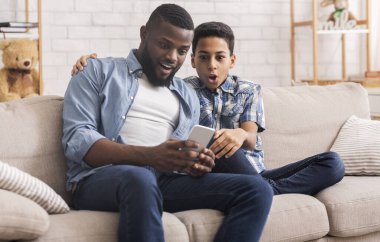 Şaşkın afro baba ve torun oğlu akıllı telefonlarıyla kanepede oturuyor.
