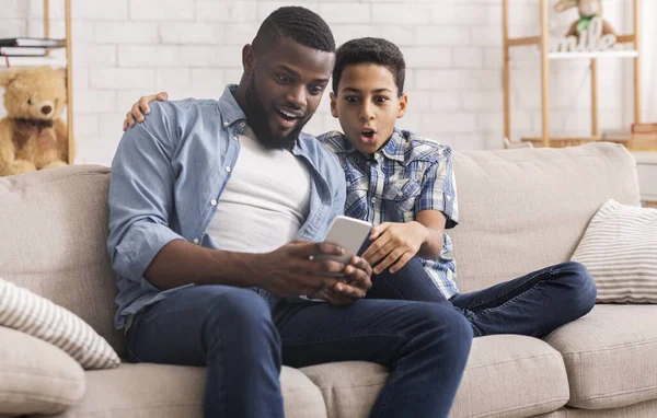 Sorprendido afro papá y preadolescente hijo sentado con teléfono inteligente en el sofá — Foto de Stock