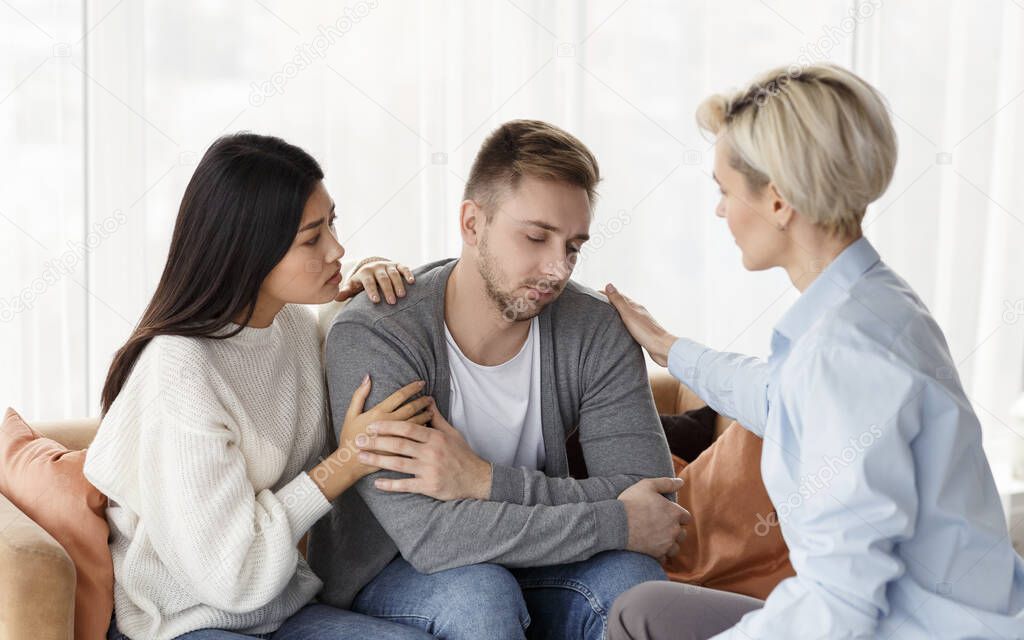 Wife And Psychologist Comforting Addicted Husband At Psychotherapy Session Indoor