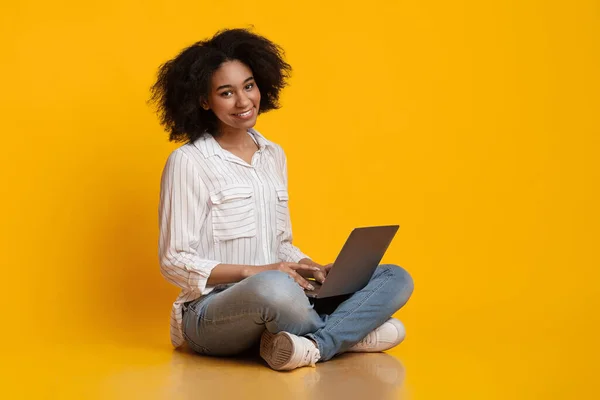 Afro kadın yerde oturuyor, dizüstü bilgisayar kullanıyor ve kameraya gülümsüyor. — Stok fotoğraf