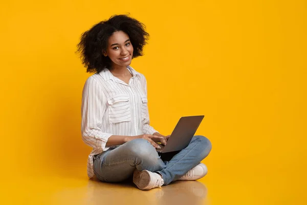 Afro mujer sentada en el suelo, usando portátil y sonriendo a la cámara — Foto de Stock