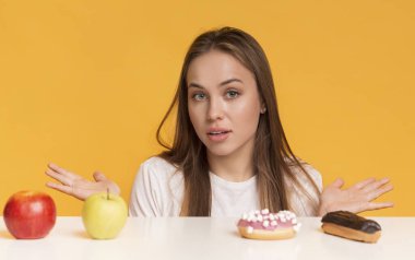 Doubtful Woman Choosing Between Healthy And Unhealthy Food, Fruits And Sweets