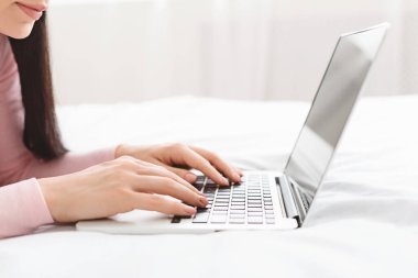 Young student typing on laptop, lying on bed at home