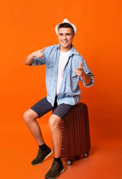 Happy young tourist sitting on suitcase and pointing at camera — Stok fotoğraf
