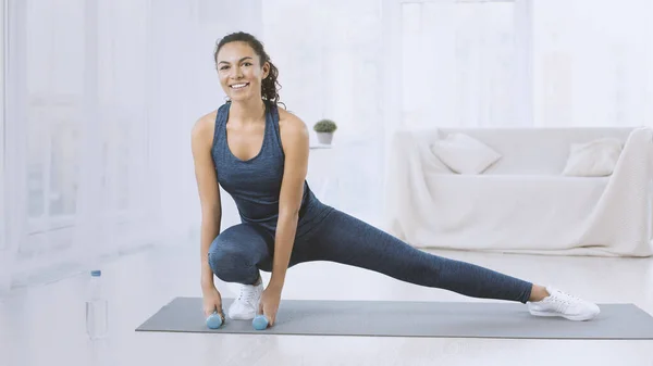 Happy Hispanic woman with dumbbells stretching on yoga mat at home — Stockfoto